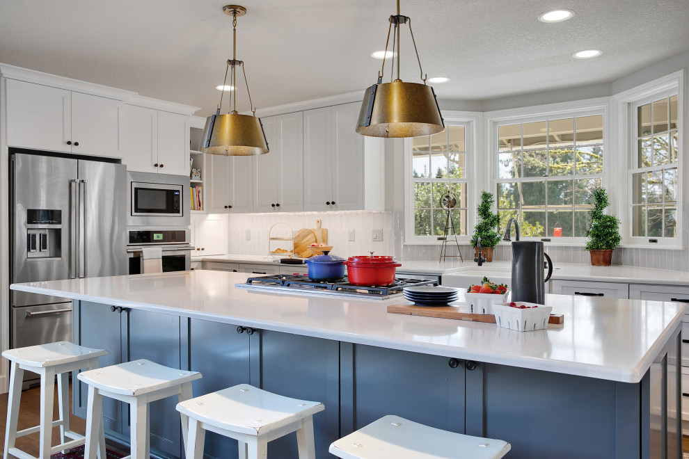 This is an example of a large farmhouse u-shaped kitchen/diner in Portland with a belfast sink, shaker cabinets, white cabinets, engineered stone countertops, grey splashback, ceramic splashback, stainless steel appliances, medium hardwood flooring, an island, brown floors and white worktops.