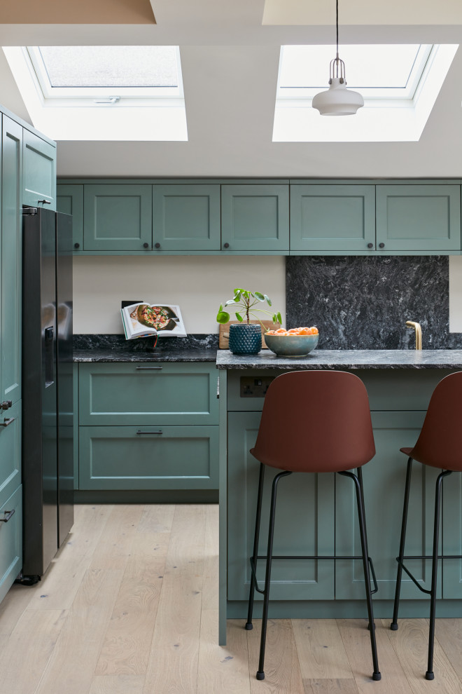 Photo of a mid-sized traditional l-shaped open plan kitchen in London with a farmhouse sink, recessed-panel cabinets, green cabinets, granite benchtops, black splashback, granite splashback, light hardwood floors, with island, black benchtop and vaulted.