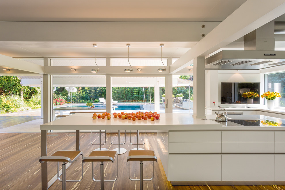 Large contemporary l-shaped open plan kitchen in Stuttgart with flat-panel cabinets, white cabinets, solid surface benchtops, with island, window splashback, white benchtop and medium hardwood floors.