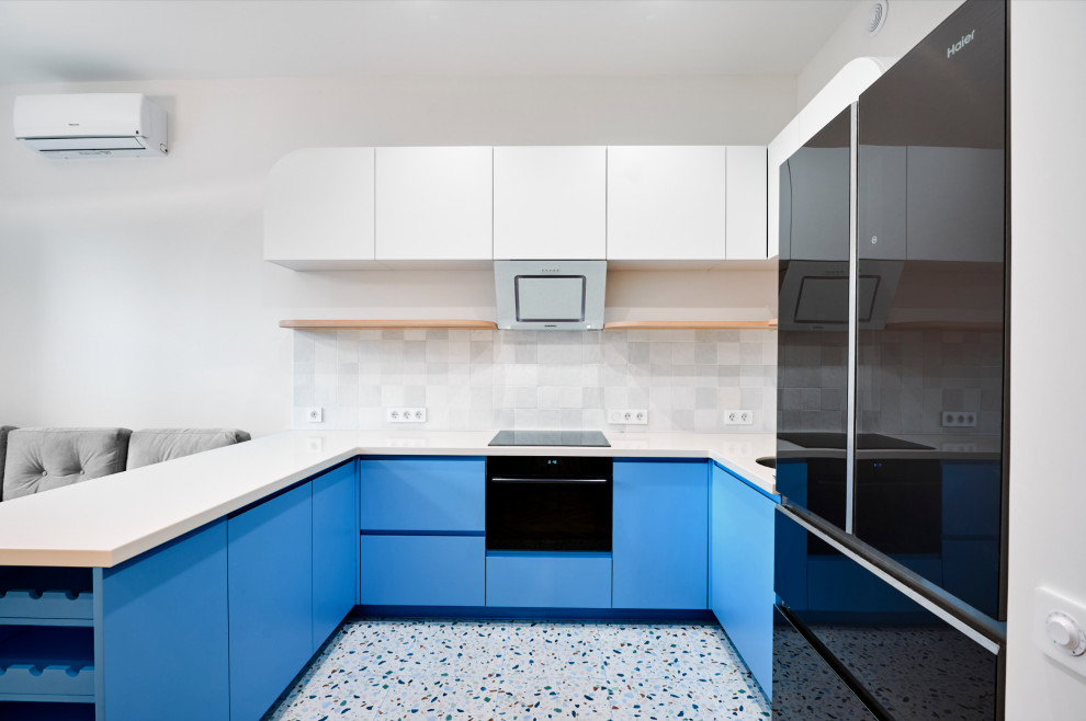 Пример оригинального дизайна: маленькая п-образная, светлая кухня-гостиная в стиле модернизм с монолитной мойкой, плоскими фасадами, синими фасадами, столешницей из акрилового камня, белым фартуком, фартуком из керамической плитки, черной техникой, полом из терраццо, полуостровом, разноцветным полом и бежевой столешницей для на участке и в саду