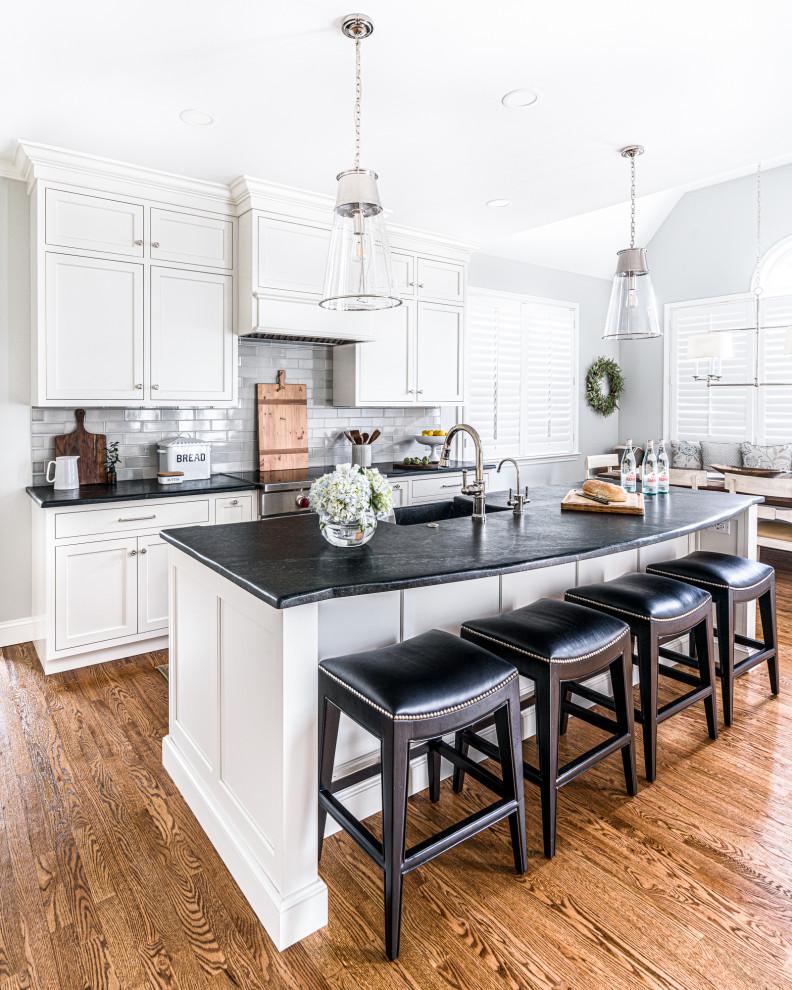 Large transitional galley medium tone wood floor and brown floor eat-in kitchen photo in Philadelphia with an undermount sink, shaker cabinets, white cabinets, gray backsplash, subway tile backsplash, stainless steel appliances, an island and black countertops