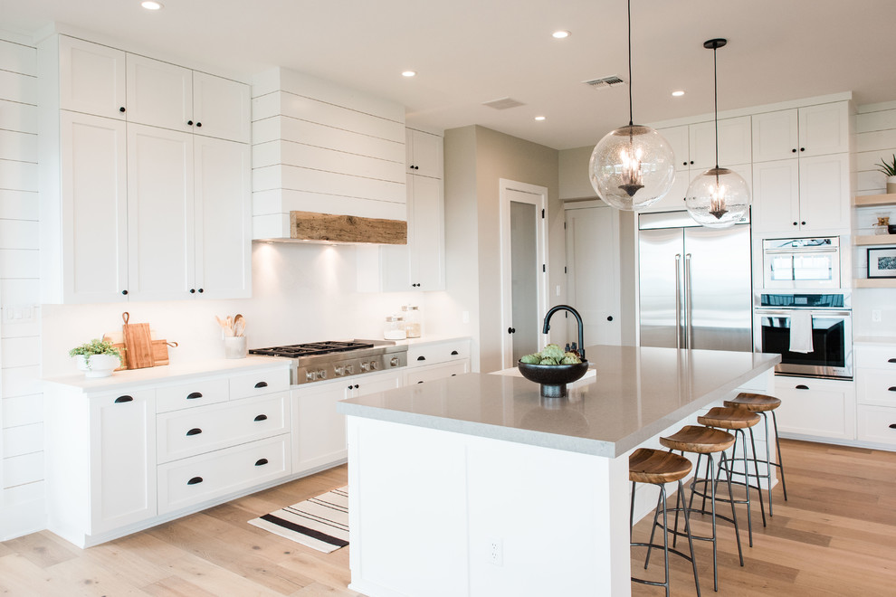 Design ideas for a large transitional u-shaped open plan kitchen in Austin with a farmhouse sink, shaker cabinets, white cabinets, quartzite benchtops, white splashback, stone slab splashback, stainless steel appliances, light hardwood floors, with island, grey benchtop and beige floor.