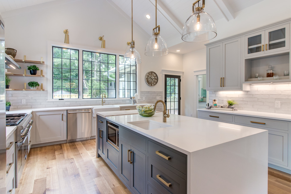 Photo of a large country u-shaped kitchen in Raleigh with a farmhouse sink, shaker cabinets, grey cabinets, grey splashback, stainless steel appliances, light hardwood floors, with island and white benchtop.