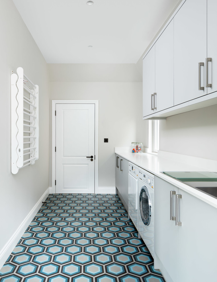 This is an example of a contemporary single-wall dedicated laundry room in London with flat-panel cabinets, grey cabinets, grey walls, a side-by-side washer and dryer, multi-coloured floor and white benchtop.