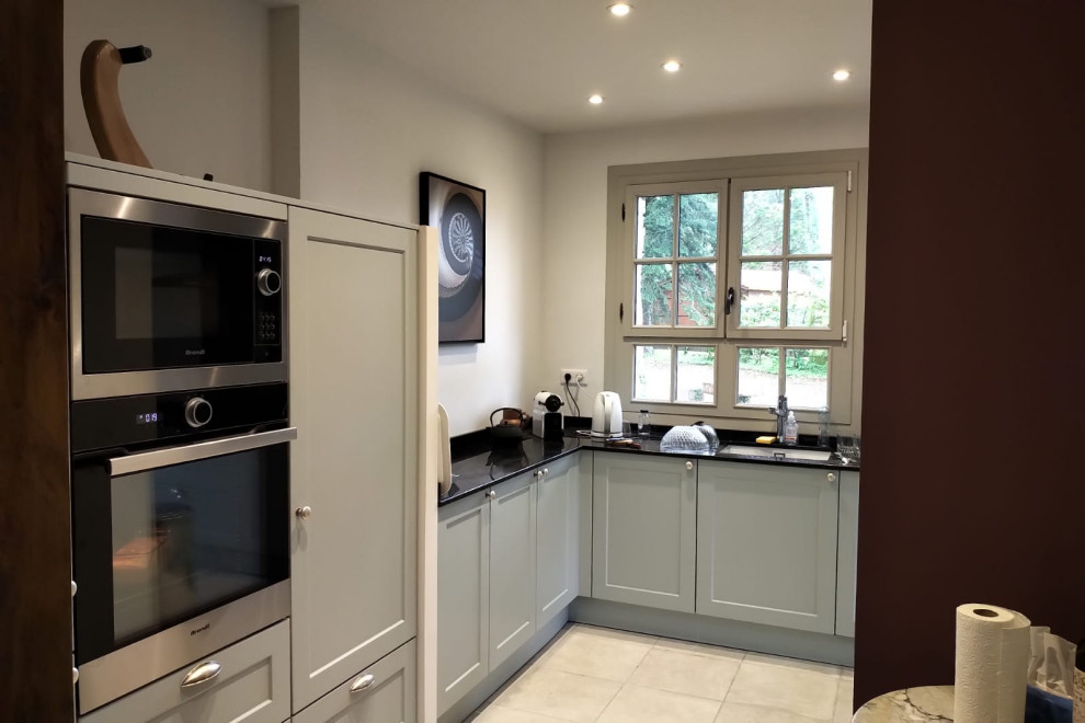 This is an example of a large transitional l-shaped open plan kitchen in Bordeaux with an undermount sink, recessed-panel cabinets, turquoise cabinets, marble benchtops, black splashback, marble splashback, black appliances, ceramic floors, white floor, black benchtop and exposed beam.