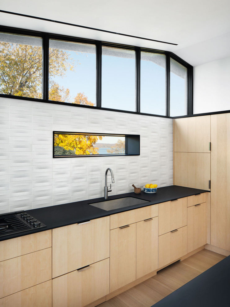 This is an example of a modern kitchen in New York with an undermount sink, flat-panel cabinets, light wood cabinets, laminate benchtops, white splashback, ceramic splashback, black appliances, light hardwood floors, with island, black benchtop and vaulted.