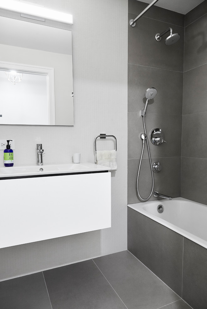 This is an example of a small contemporary master bathroom in New York with flat-panel cabinets, white cabinets, a drop-in tub, a shower/bathtub combo, a one-piece toilet, gray tile, porcelain tile, white walls, porcelain floors, a wall-mount sink, grey floor, a shower curtain and solid surface benchtops.