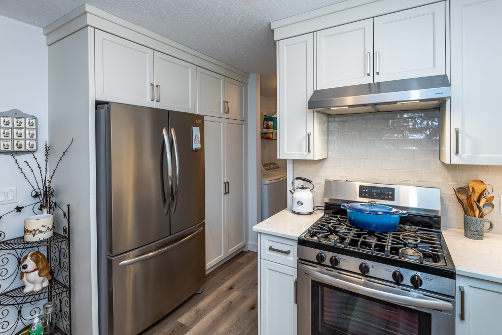 This is an example of a transitional u-shaped kitchen in Toronto with an undermount sink, white cabinets, quartz benchtops, white splashback, ceramic splashback, stainless steel appliances, vinyl floors, with island, brown floor and white benchtop.