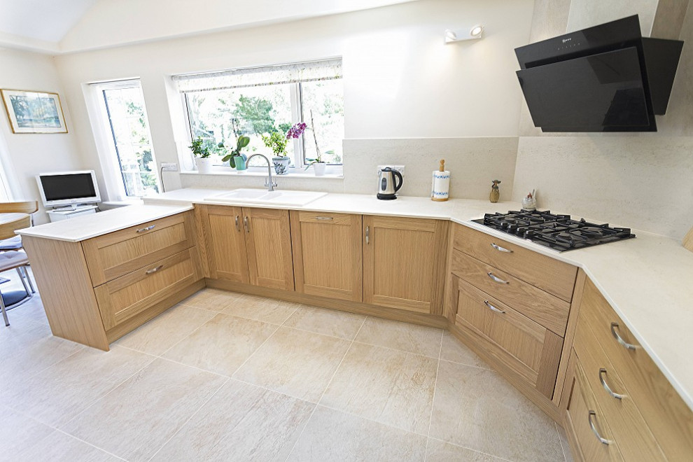 Photo of a small traditional u-shaped eat-in kitchen in Oxfordshire with a single-bowl sink, flat-panel cabinets, medium wood cabinets, white splashback, black appliances, porcelain floors, no island, grey floor and white benchtop.