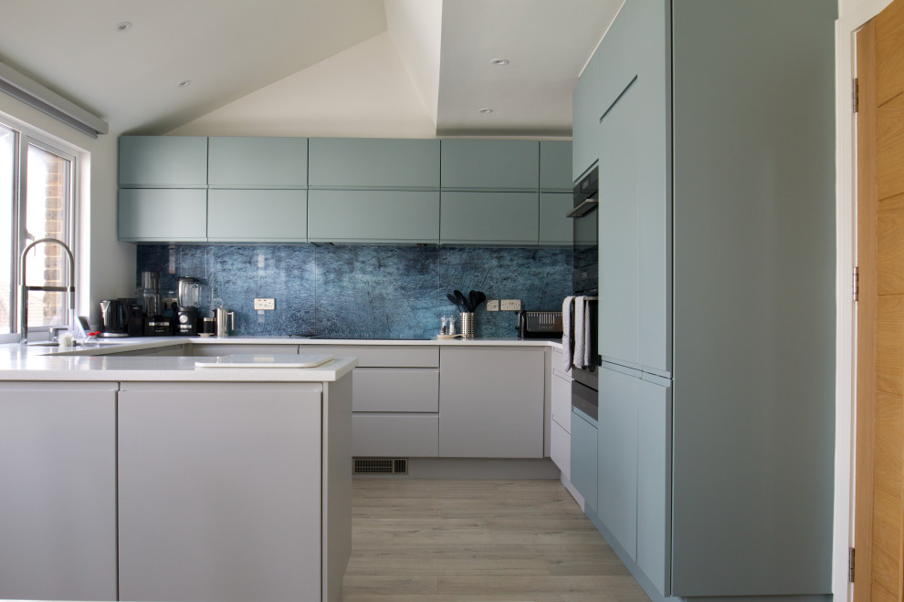 Inspiration for a mid-sized beach style u-shaped eat-in kitchen in Hampshire with an undermount sink, flat-panel cabinets, blue cabinets, solid surface benchtops, blue splashback, glass sheet splashback, black appliances, laminate floors, no island, beige floor, white benchtop and vaulted.