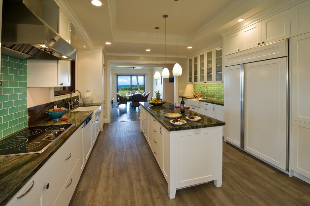 Design ideas for a tropical kitchen in Hawaii with a farmhouse sink, shaker cabinets, white cabinets, granite benchtops, green splashback, glass tile splashback, panelled appliances, vinyl floors and with island.
