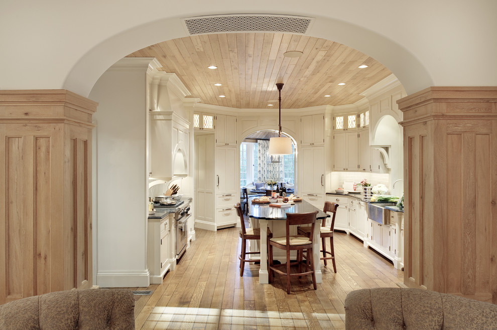 Mid-sized traditional u-shaped eat-in kitchen in New York with a farmhouse sink, white cabinets, white splashback, stainless steel appliances, medium hardwood floors, with island and recessed-panel cabinets.