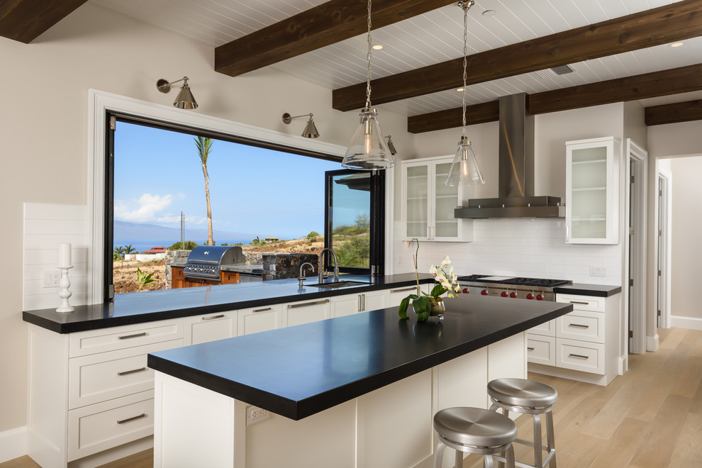 Photo of a transitional l-shaped kitchen in Hawaii with an undermount sink, granite benchtops, white splashback, porcelain splashback, stainless steel appliances, light hardwood floors, with island and glass-front cabinets.