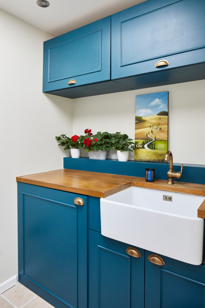 This is an example of a small traditional single-wall dedicated laundry room in London with a farmhouse sink, shaker cabinets, blue cabinets, wood benchtops, beige walls, travertine floors, beige floor and brown benchtop.