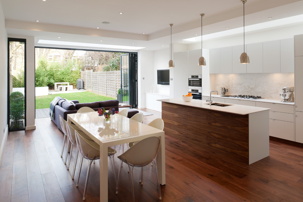 Design ideas for a mid-sized contemporary single-wall open plan kitchen in London with flat-panel cabinets, white cabinets, solid surface benchtops, white splashback, stone slab splashback, medium hardwood floors, with island, a double-bowl sink, stainless steel appliances and brown floor.