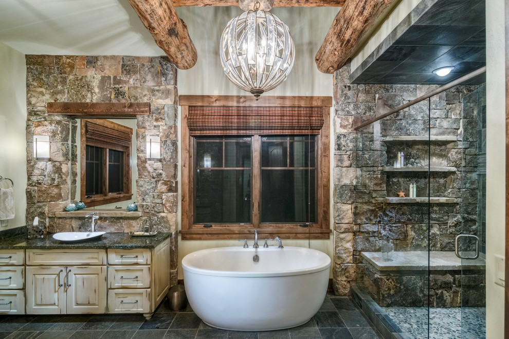 Inspiration for a large rustic master travertine floor and gray floor freestanding bathtub remodel with distressed cabinets, beige walls, a pedestal sink, granite countertops, a hinged shower door and black countertops
