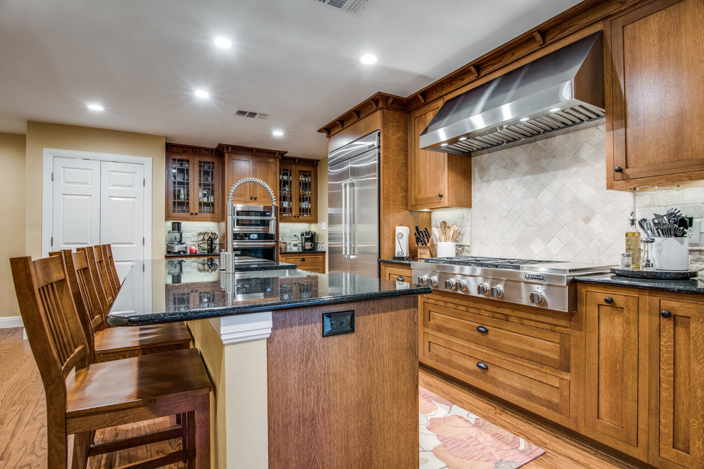 Large arts and crafts l-shaped eat-in kitchen in Austin with a farmhouse sink, shaker cabinets, medium wood cabinets, granite benchtops, beige splashback, ceramic splashback, stainless steel appliances, light hardwood floors, with island, brown floor and black benchtop.