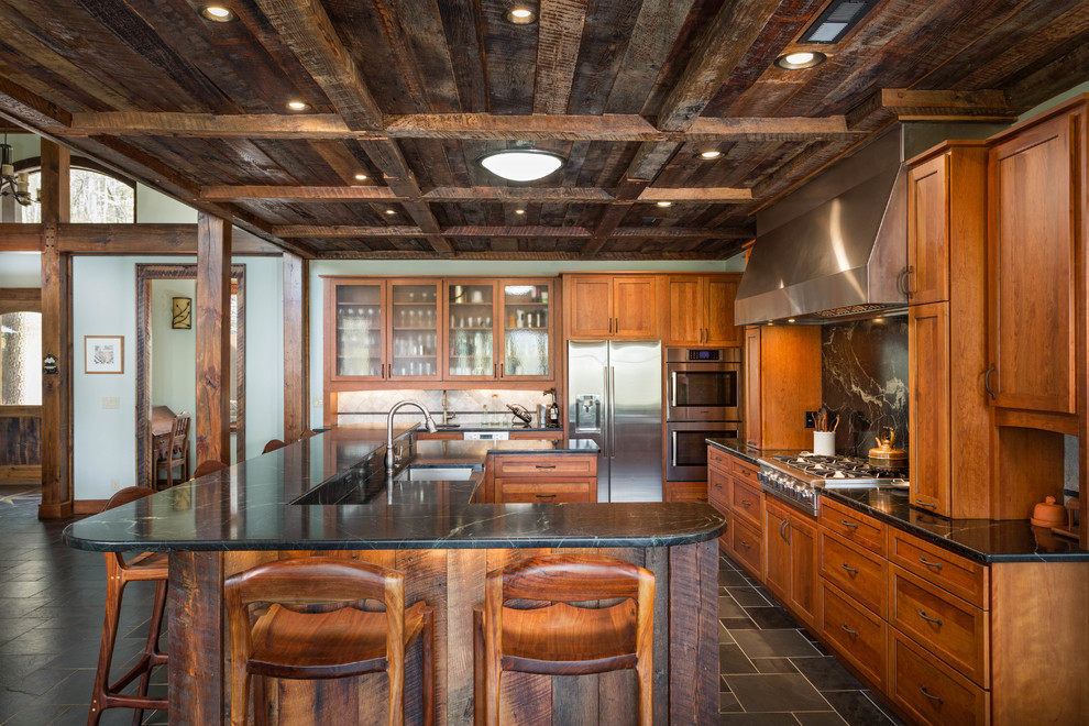 Large mountain style u-shaped porcelain tile and gray floor eat-in kitchen photo with a farmhouse sink, shaker cabinets, medium tone wood cabinets, soapstone countertops, stone slab backsplash, stainless steel appliances and an island