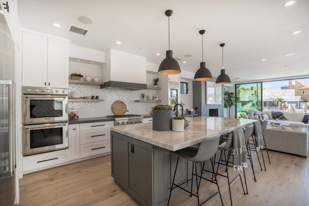 This is an example of a contemporary l-shaped kitchen in Orange County with shaker cabinets, white cabinets, grey splashback, stainless steel appliances, medium hardwood floors, with island, brown floor and grey benchtop.