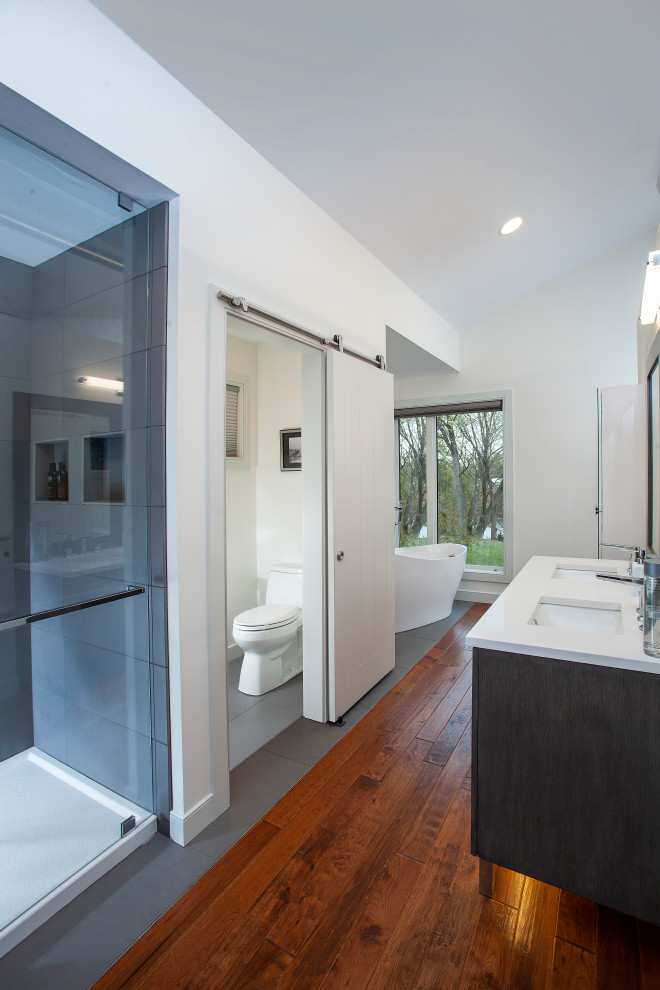Design ideas for a mid-sized modern master bathroom in Other with flat-panel cabinets, dark wood cabinets, a freestanding tub, a double shower, a one-piece toilet, gray tile, ceramic tile, white walls, ceramic floors, an undermount sink, engineered quartz benchtops, grey floor, a hinged shower door and white benchtops.