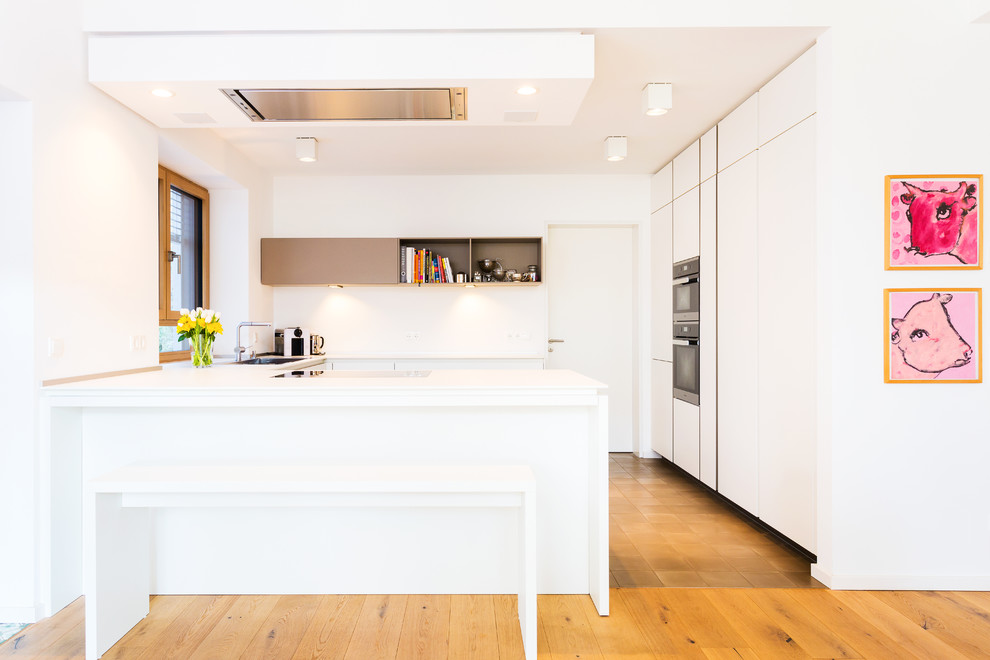 Photo of a mid-sized contemporary u-shaped open plan kitchen in Frankfurt with flat-panel cabinets, white cabinets, white splashback, stainless steel appliances, light hardwood floors, a peninsula, glass sheet splashback and a drop-in sink.