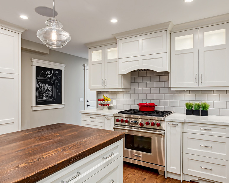 Mid-sized transitional l-shaped light wood floor and beige floor enclosed kitchen photo in Calgary with a farmhouse sink, recessed-panel cabinets, white cabinets, quartz countertops, white backsplash, porcelain backsplash, stainless steel appliances and an island