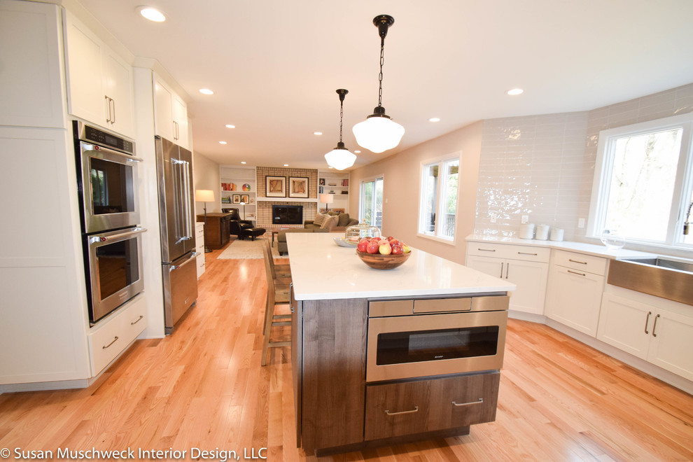 Inspiration for a mid-sized transitional light wood floor and brown floor kitchen remodel with a farmhouse sink, shaker cabinets, white cabinets, quartz countertops, gray backsplash, ceramic backsplash, stainless steel appliances, an island and white countertops