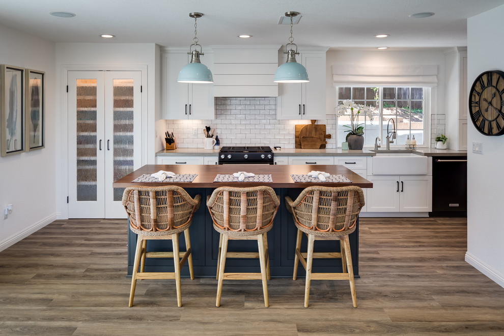Mid-sized country eat-in kitchen in San Diego with a farmhouse sink, shaker cabinets, white cabinets, white splashback, subway tile splashback, black appliances, medium hardwood floors, with island, brown floor, wood benchtops and brown benchtop.