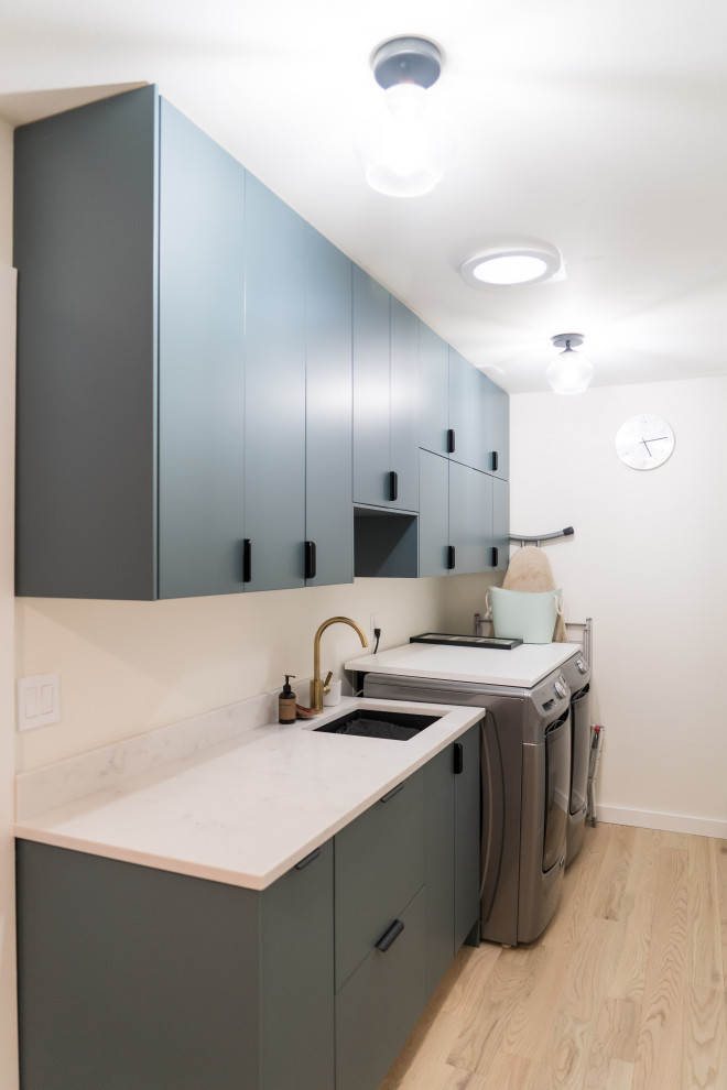 Photo of a small scandinavian galley utility room in Seattle with an undermount sink, flat-panel cabinets, green cabinets, quartz benchtops, white splashback, engineered quartz splashback, beige walls, light hardwood floors, a side-by-side washer and dryer, beige floor and white benchtop.