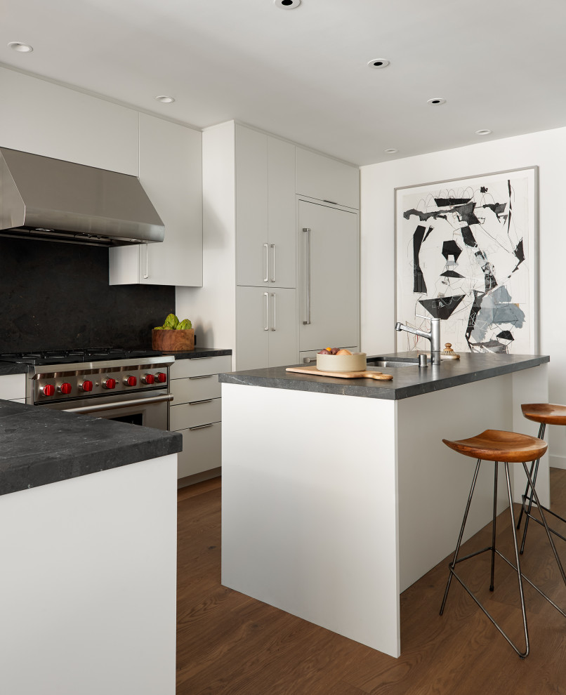 Example of an urban l-shaped medium tone wood floor and brown floor kitchen design in San Francisco with an undermount sink, flat-panel cabinets, white cabinets, black backsplash, stone slab backsplash, paneled appliances, an island and black countertops