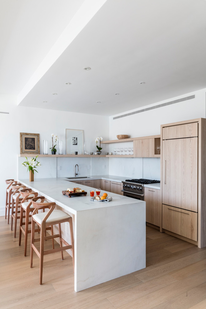 This is an example of a modern galley open plan kitchen in New York with an undermount sink, flat-panel cabinets, light wood cabinets, white splashback, stainless steel appliances, light hardwood floors, a peninsula and beige floor.