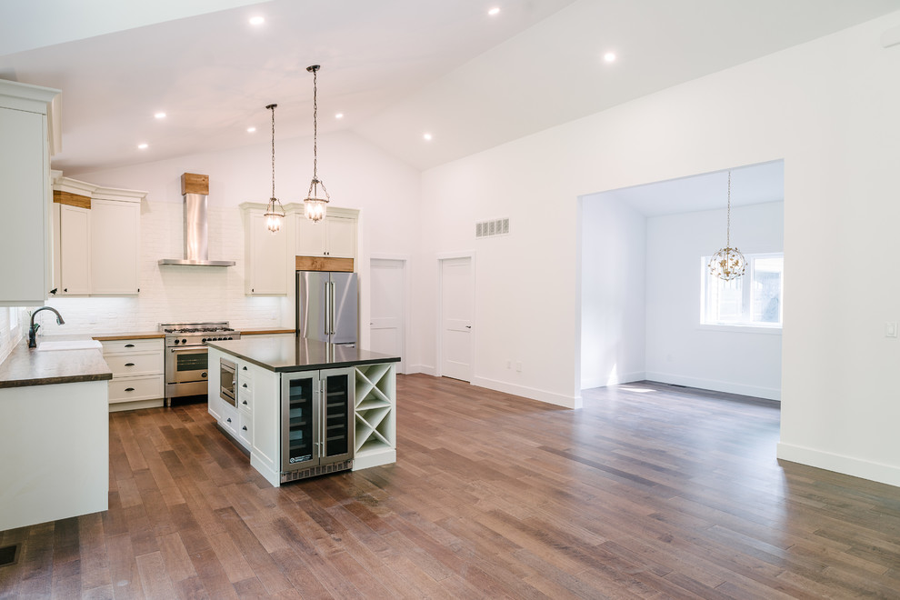 This is an example of a mid-sized modern l-shaped eat-in kitchen with a farmhouse sink, shaker cabinets, white cabinets, wood benchtops, white splashback, brick splashback, stainless steel appliances, with island and beige benchtop.