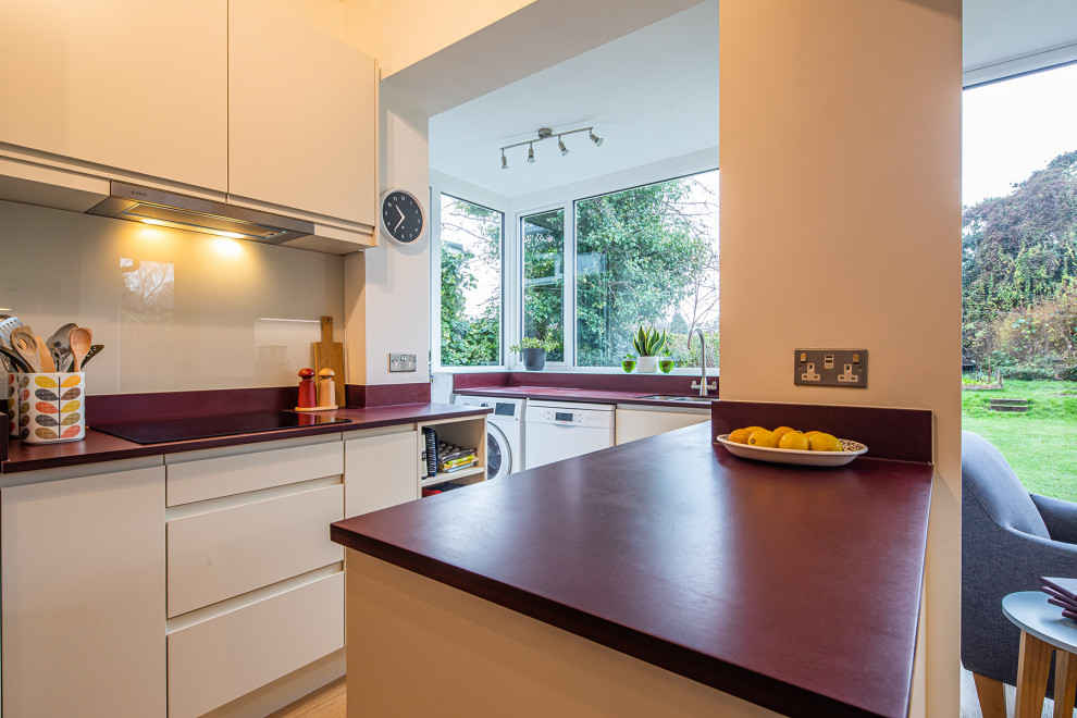 This is an example of a small contemporary u-shaped eat-in kitchen in Other with flat-panel cabinets, white cabinets, white splashback, glass sheet splashback, stainless steel appliances, no island, brown floor and red benchtop.