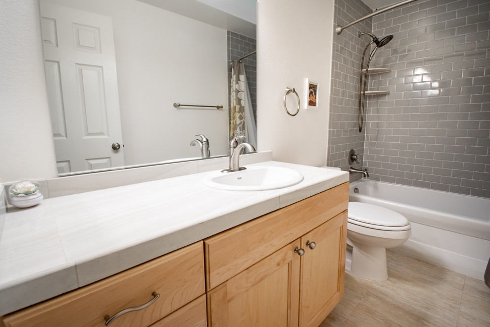 This is an example of a mid-sized transitional kids bathroom in Seattle with recessed-panel cabinets, light wood cabinets, an alcove tub, a shower/bathtub combo, a two-piece toilet, gray tile, ceramic tile, grey walls, vinyl floors, a drop-in sink, tile benchtops, beige floor, a shower curtain, grey benchtops, a single vanity and a built-in vanity.