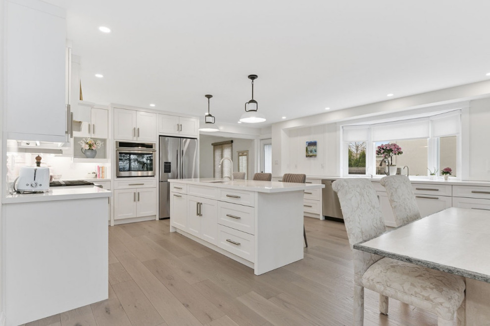 Exempel på ett stort vit vitt kök och matrum, med skåp i shakerstil, vita skåp, marmorbänkskiva, vitt stänkskydd, en köksö, rostfria vitvaror, ljust trägolv och brunt golv