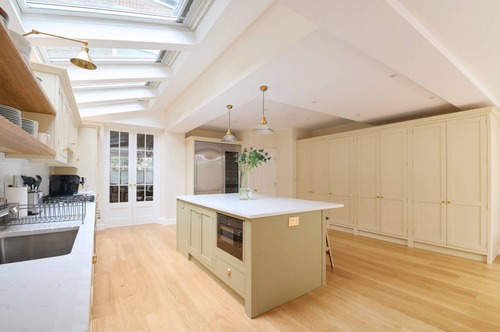 This is an example of a mid-sized traditional single-wall open plan kitchen in London with an undermount sink, shaker cabinets, beige cabinets, quartzite benchtops, white splashback, engineered quartz splashback, stainless steel appliances, light hardwood floors, with island, brown floor, white benchtop and vaulted.