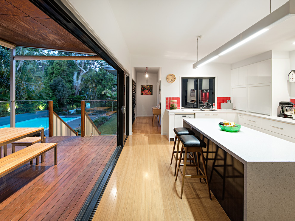 Photo of a large contemporary l-shaped eat-in kitchen in Brisbane with flat-panel cabinets, white cabinets, quartz benchtops, red splashback, ceramic splashback, light hardwood floors, with island, beige floor, white benchtop and a drop-in sink.