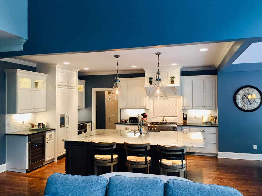 Large transitional l-shaped medium tone wood floor and brown floor open concept kitchen photo in Charlotte with a farmhouse sink, raised-panel cabinets, white cabinets, granite countertops, white backsplash, porcelain backsplash, stainless steel appliances, an island and white countertops