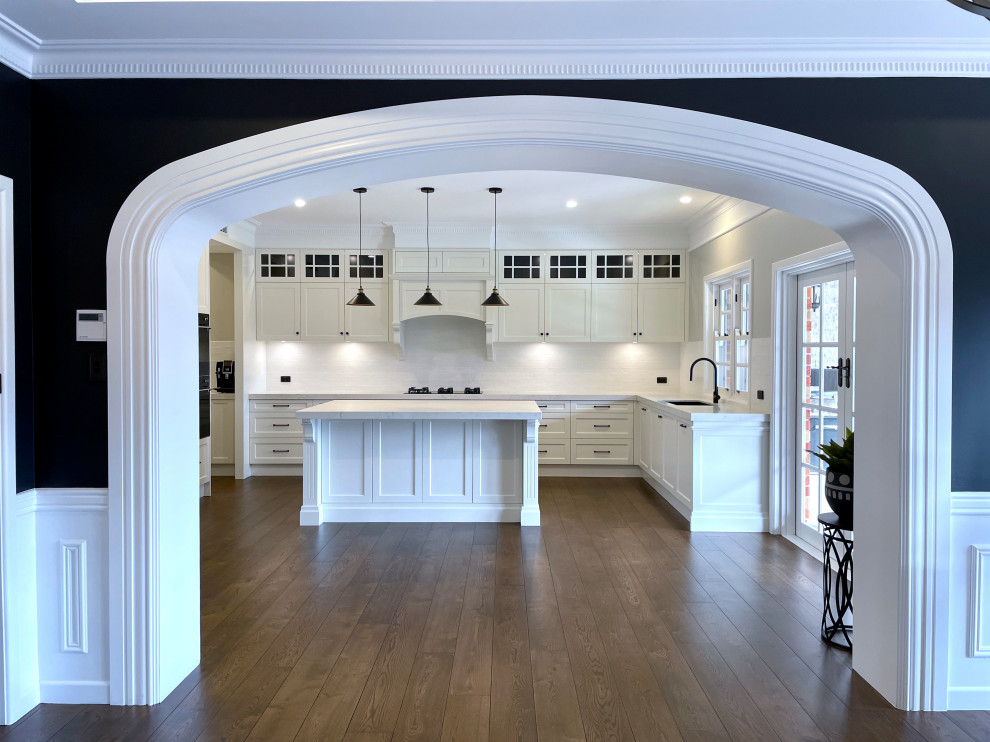 Photo of an expansive traditional u-shaped open plan kitchen in Sydney with an undermount sink, recessed-panel cabinets, white cabinets, quartz benchtops, white splashback, subway tile splashback, black appliances, medium hardwood floors, with island, brown floor and white benchtop.