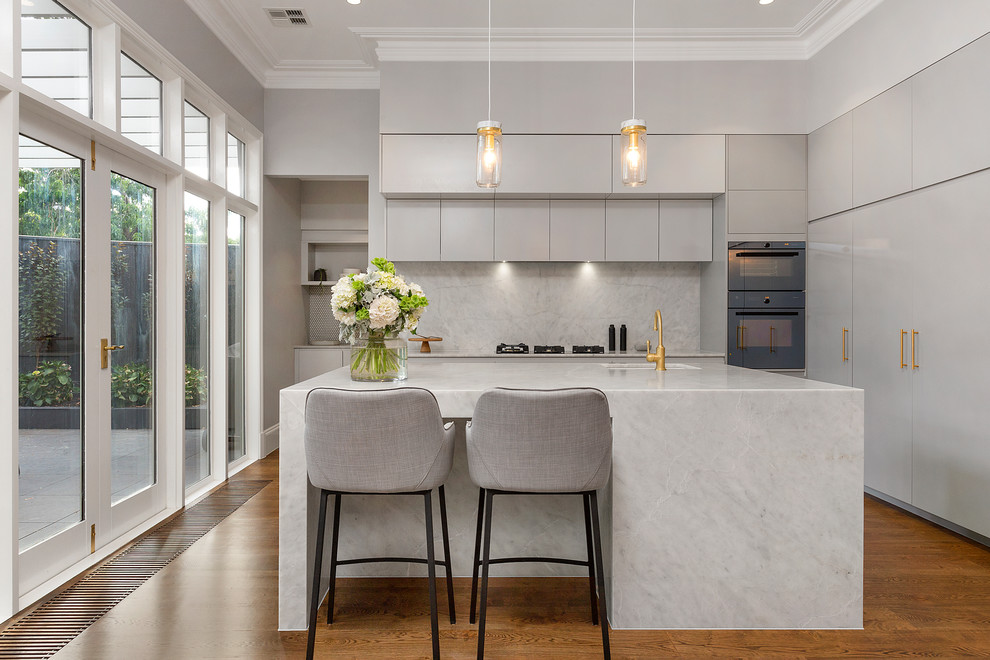 Inspiration for a contemporary l-shaped kitchen in Melbourne with an undermount sink, flat-panel cabinets, grey cabinets, white splashback, medium hardwood floors, with island, brown floor, marble splashback and black appliances.