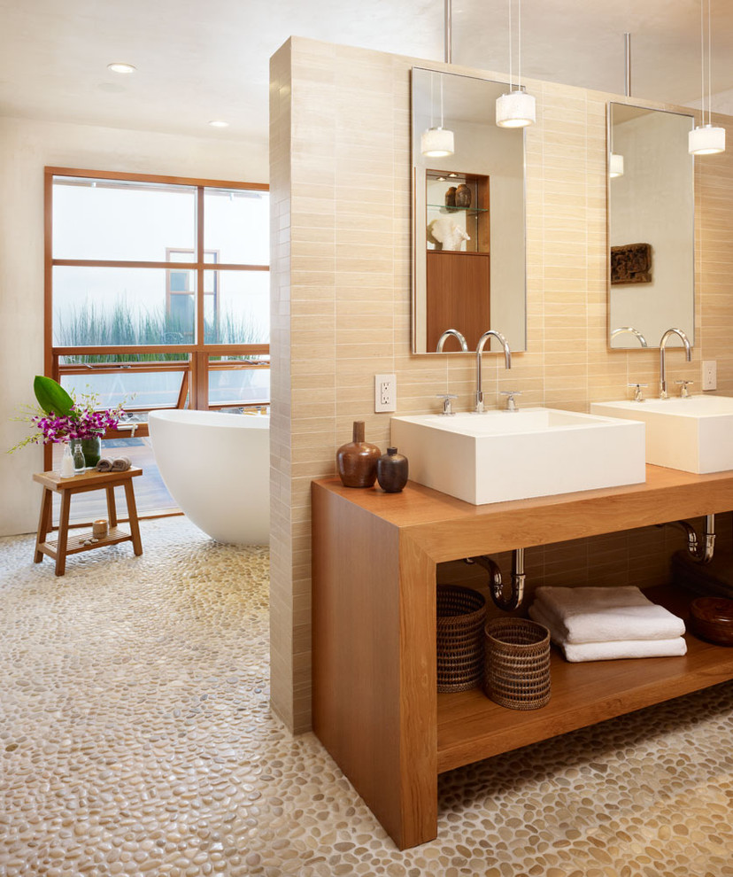 This is an example of a mid-sized tropical master wet room bathroom in Los Angeles with a freestanding tub, a vessel sink, pebble tile, pebble tile floors, open cabinets, dark wood cabinets, a wall-mount toilet, beige tile, beige walls, wood benchtops, multi-coloured floor, an open shower and brown benchtops.