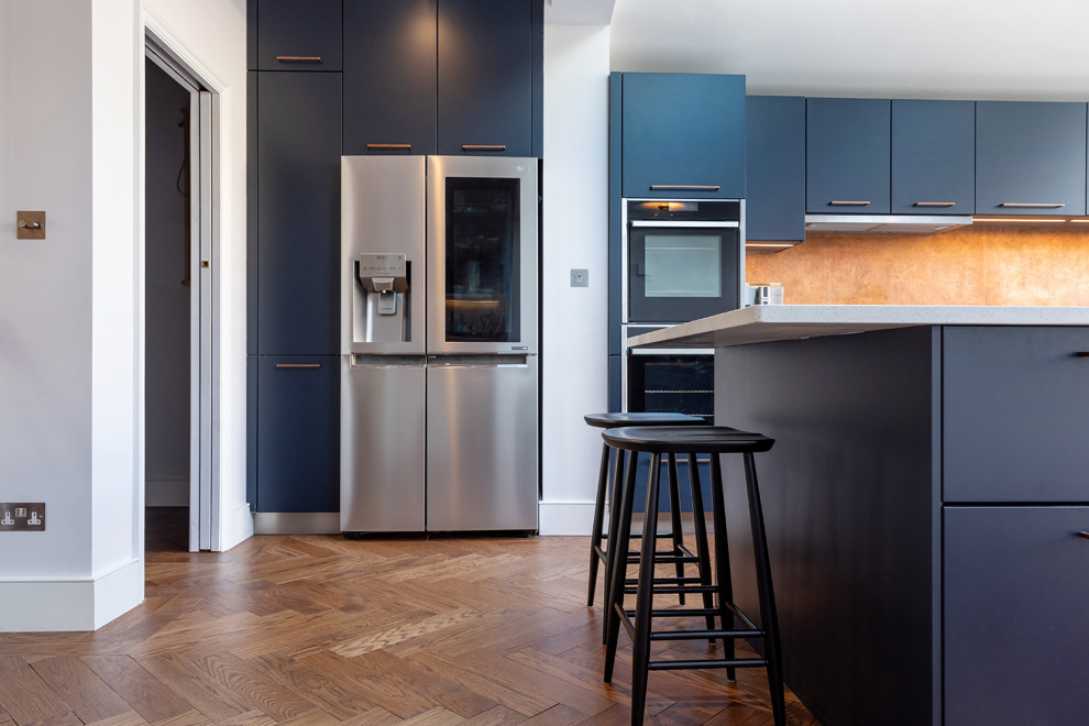 Photo of a large contemporary single-wall eat-in kitchen in London with an undermount sink, flat-panel cabinets, blue cabinets, quartzite benchtops, metallic splashback, black appliances, medium hardwood floors, with island, brown floor and white benchtop.