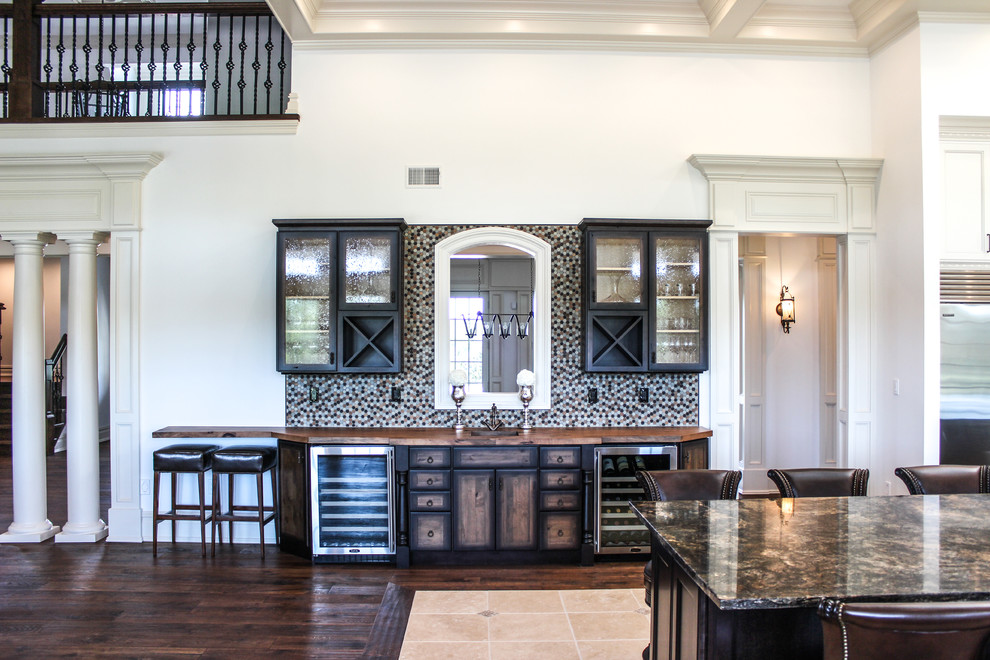 Photo of a mid-sized traditional single-wall seated home bar in New York with a drop-in sink, wood benchtops, brown splashback, medium hardwood floors, dark wood cabinets, ceramic splashback, glass-front cabinets and brown floor.