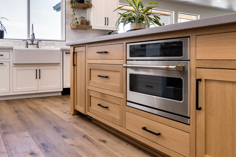 This is an example of a large country kitchen in Other with a farmhouse sink, shaker cabinets, light wood cabinets, quartz benchtops, white splashback, subway tile splashback, stainless steel appliances, vinyl floors, multiple islands, brown floor and grey benchtop.