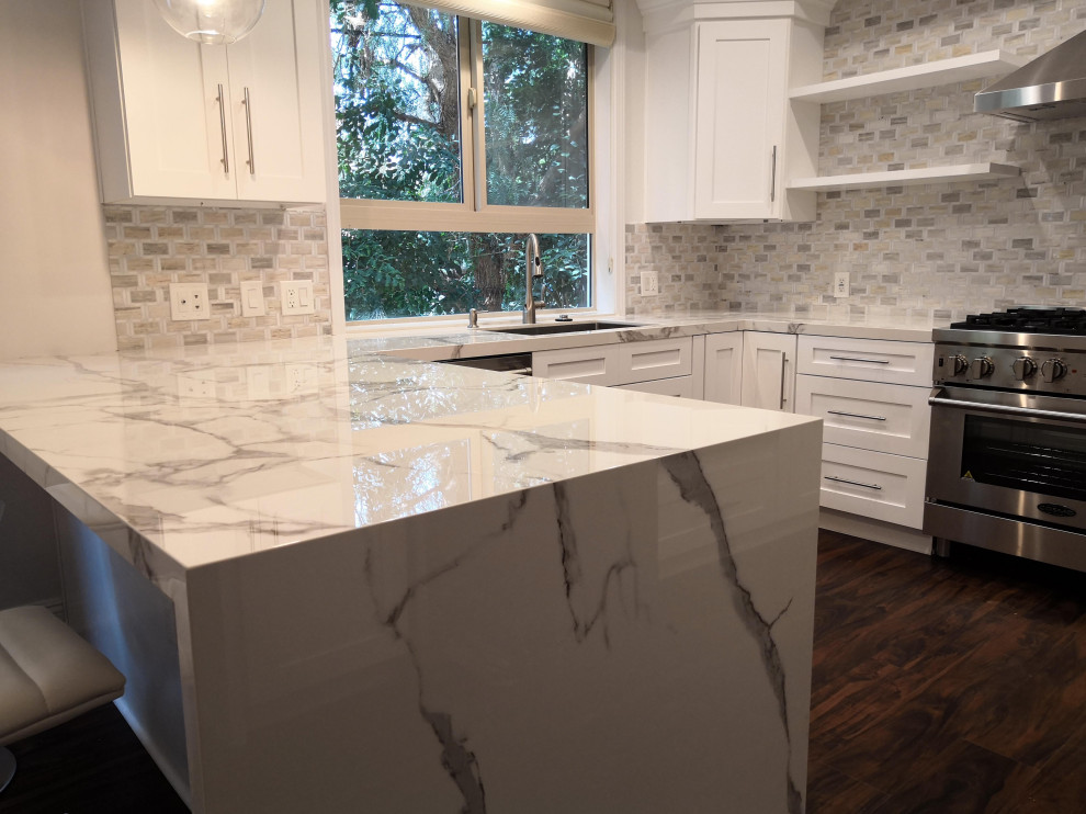 Photo of a mid-sized transitional u-shaped eat-in kitchen in Los Angeles with a single-bowl sink, recessed-panel cabinets, white cabinets, solid surface benchtops, white splashback, marble splashback, stainless steel appliances, vinyl floors, a peninsula, brown floor and white benchtop.