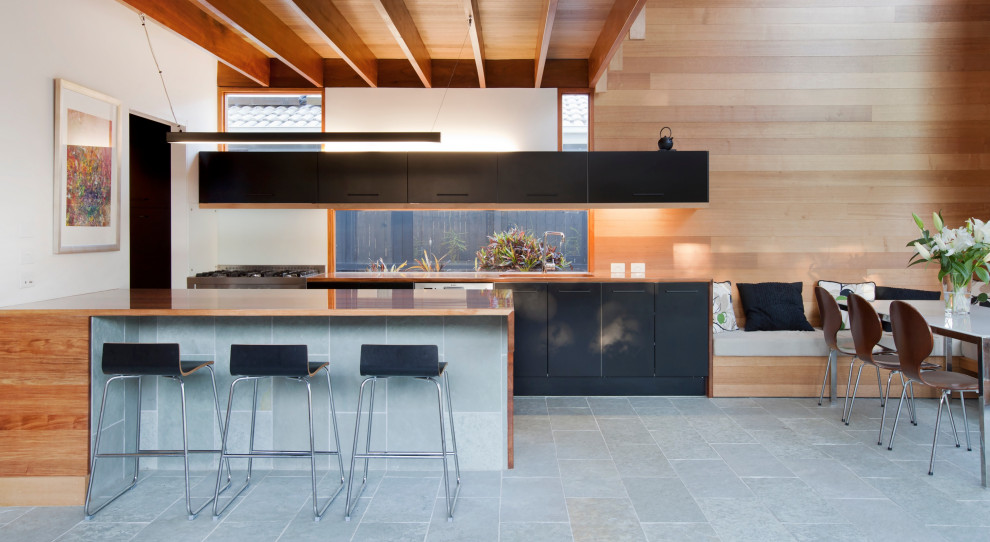 This is an example of a mid-sized contemporary galley open plan kitchen in Brisbane with a double-bowl sink, recessed-panel cabinets, black cabinets, wood benchtops, white splashback, window splashback, stainless steel appliances, slate floors, with island, grey floor, brown benchtop and exposed beam.