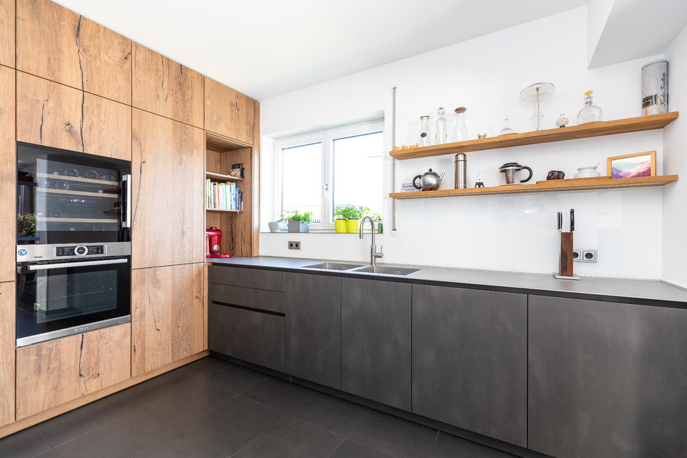 Inspiration for a large contemporary l-shaped separate kitchen in Frankfurt with an integrated sink, flat-panel cabinets, light wood cabinets, stainless steel benchtops, white splashback, glass sheet splashback, black appliances, no island, black floor and grey benchtop.