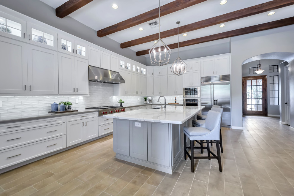 Inspiration for a large transitional u-shaped porcelain tile, beige floor and exposed beam enclosed kitchen remodel in Phoenix with a farmhouse sink, shaker cabinets, white cabinets, quartz countertops, white backsplash, glass tile backsplash, stainless steel appliances, an island and white countertops