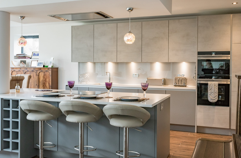 Photo of a mid-sized modern l-shaped open plan kitchen in Surrey with an integrated sink, flat-panel cabinets, grey cabinets, quartzite benchtops, grey splashback, glass sheet splashback, stainless steel appliances, medium hardwood floors, with island and brown floor.