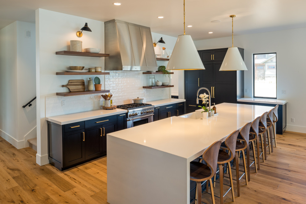 Photo of a transitional kitchen in Seattle with a farmhouse sink, shaker cabinets, black cabinets, quartz benchtops, white splashback, subway tile splashback, stainless steel appliances, medium hardwood floors, with island and white benchtop.